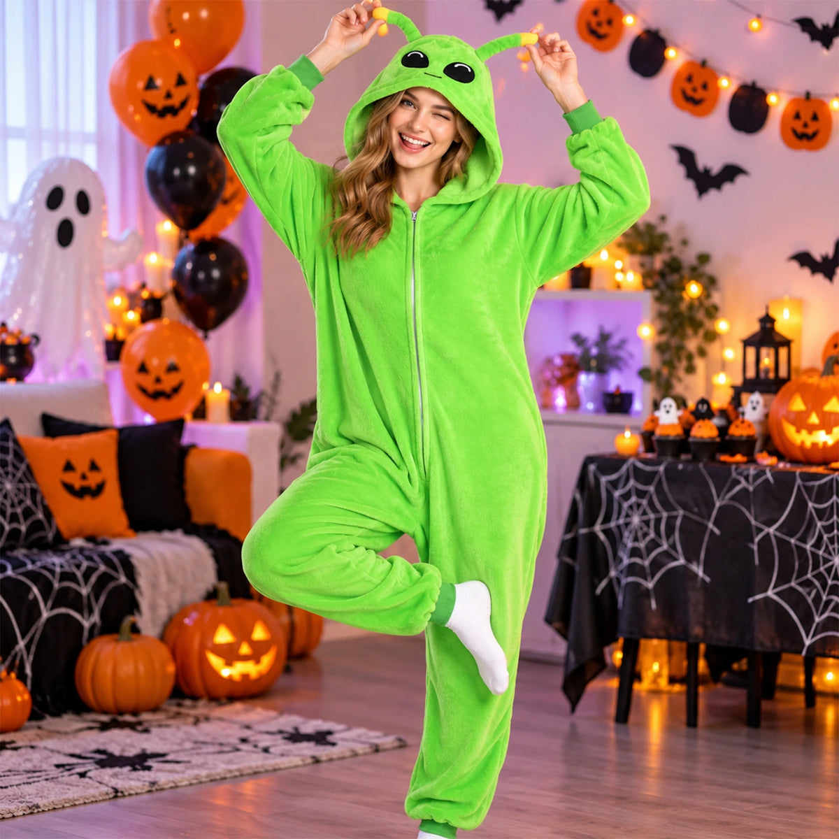 A woman in the green alien costume holds its antennae beside pumpkins and Halloween party decorations.