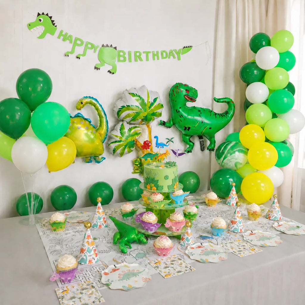 Dinosaur birthday party table and balloon setup with paper plates, party hats, cake stand and green dinosaur decorations.