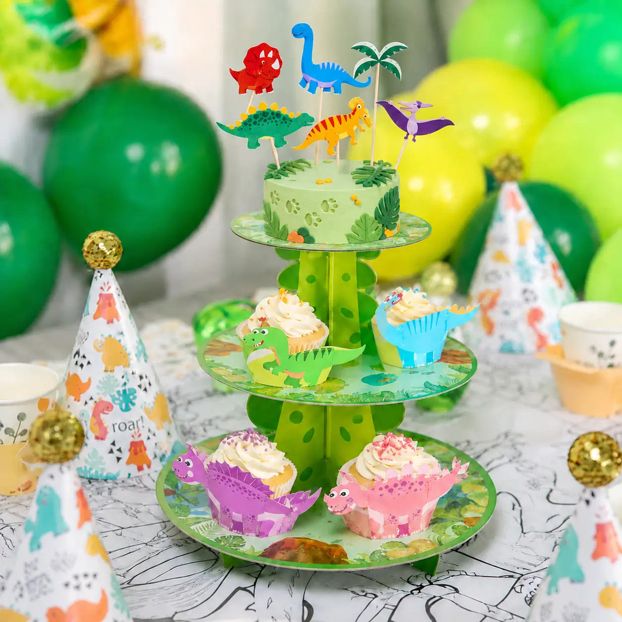 Dinosaur cake table display with colorful cake toppers, cupcake wrappers, a three-tier stand and green birthday balloons.