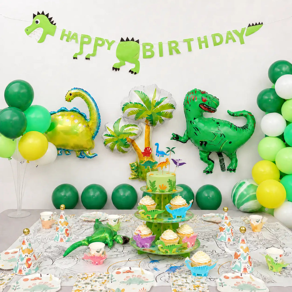 Dinosaur party decorations kit displayed with coordinated tableware, cupcakes, party hats, foil balloons and a birthday banner.