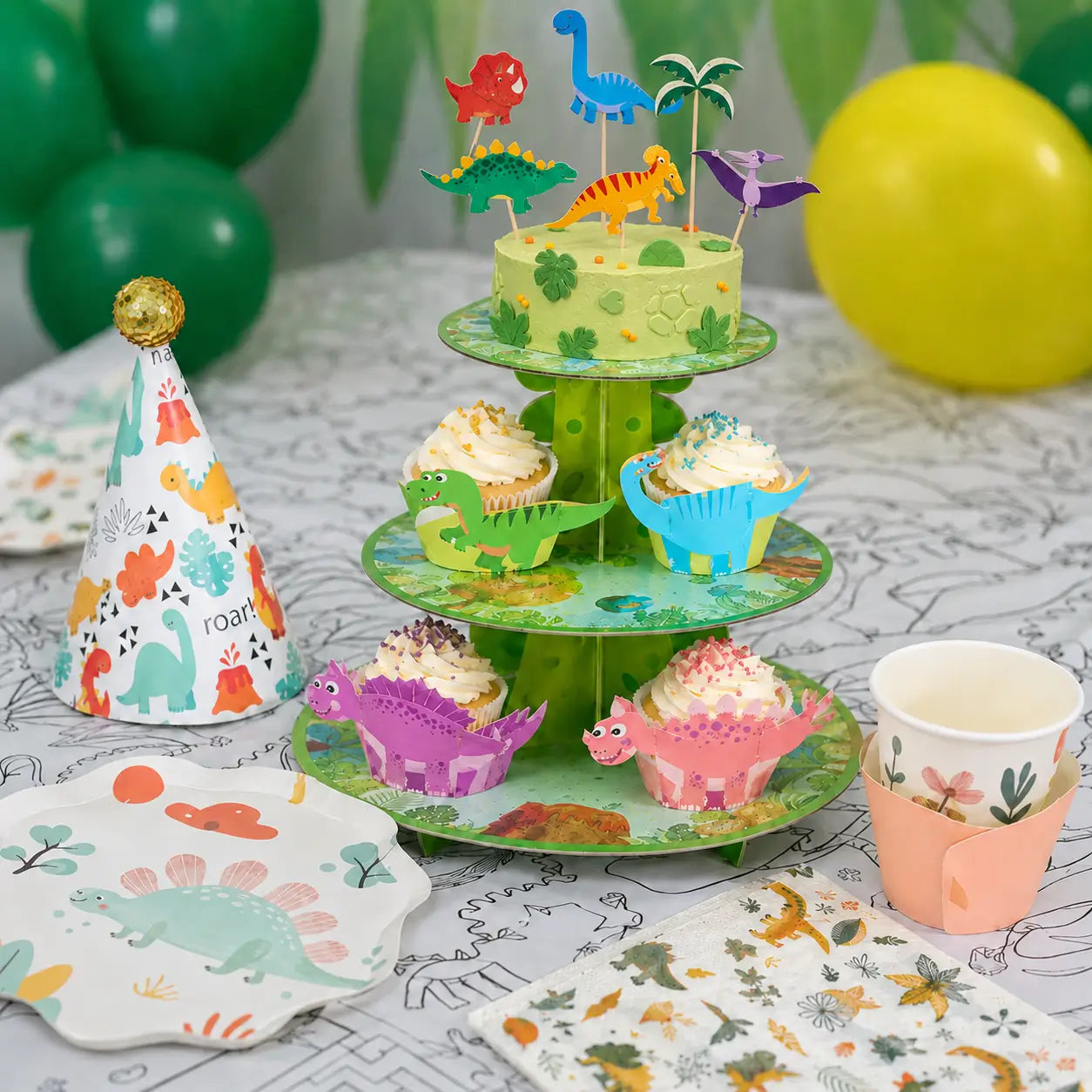 Close-up of dinosaur party tableware with a green cake stand, dinosaur cupcake wrappers, cake toppers, paper plate and cup.
