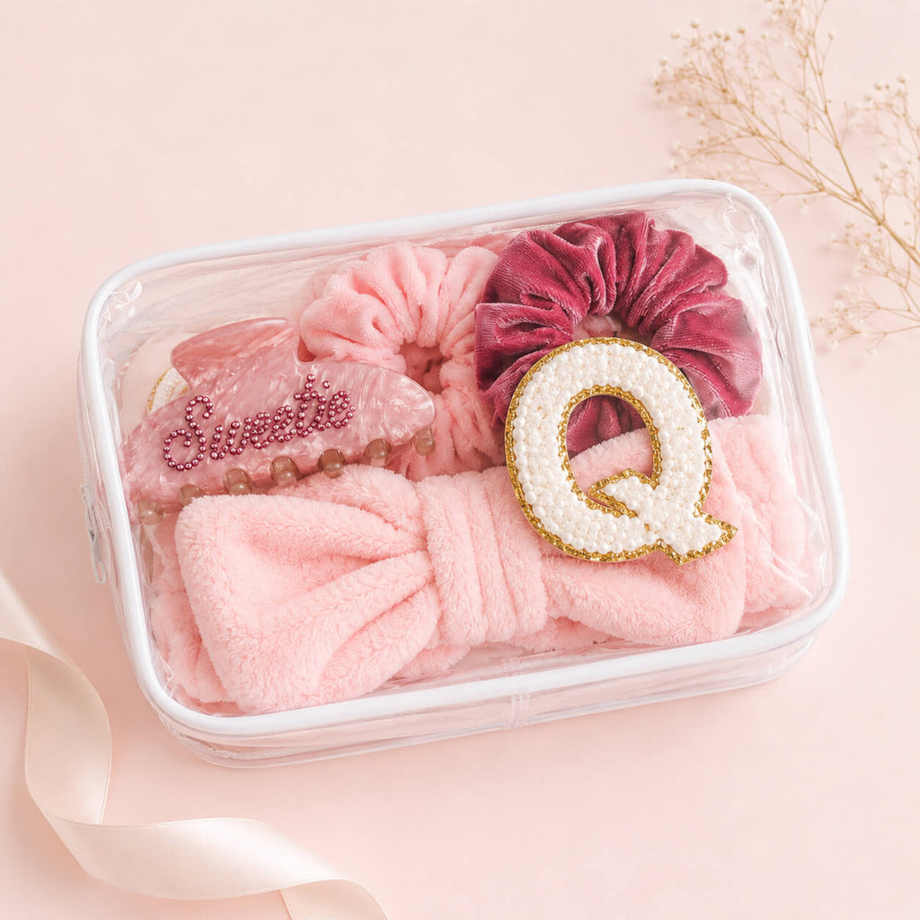 Clear cosmetic pouch filled with pink spa party accessories including fluffy headband, velvet scrunchies, pink hair clip, and chenille letter Q patch on soft pastel background