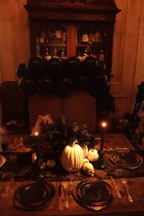 Gothic Halloween dinner party table with black balloon garland, white pumpkins, skull decor, candle holders and black-and-gold plates
