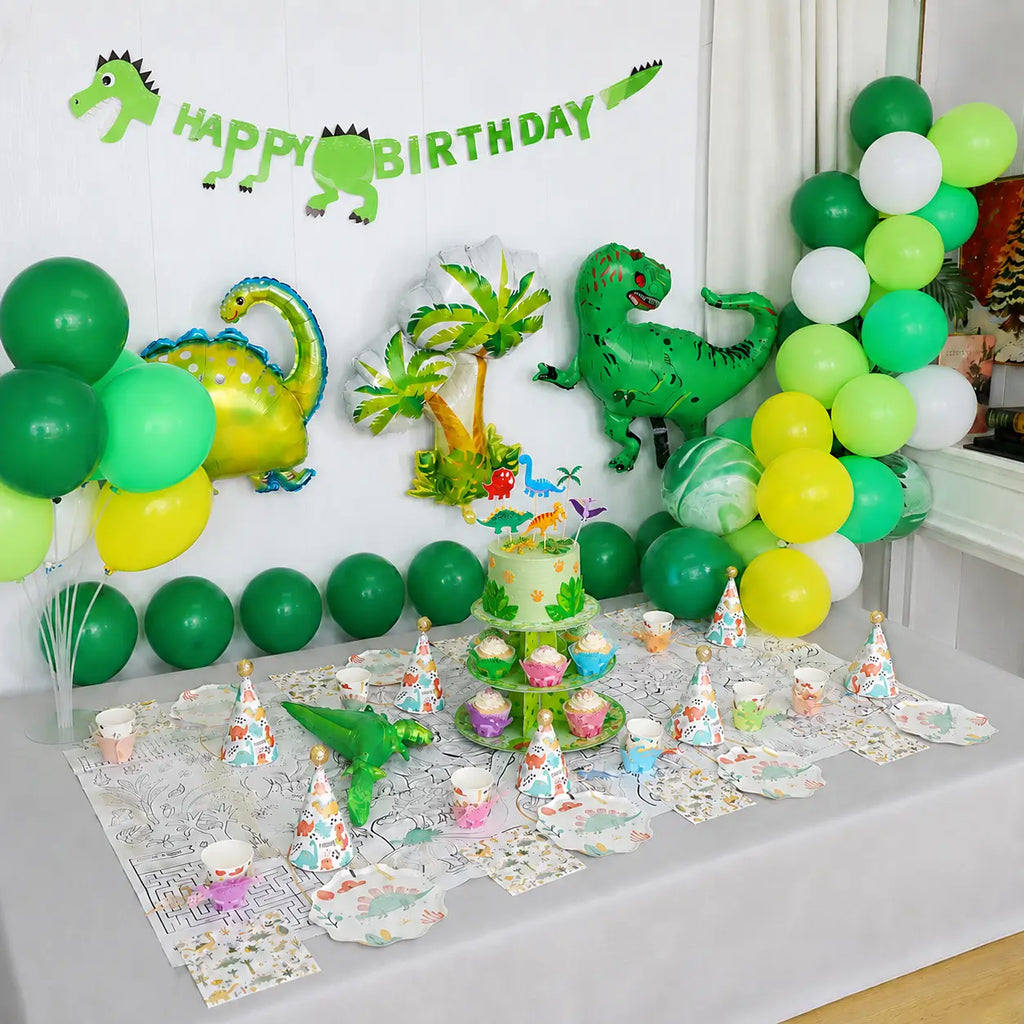 Green dinosaur party decorations for kids with a balloon backdrop, dinosaur tableware, party hats and a decorated cake table.