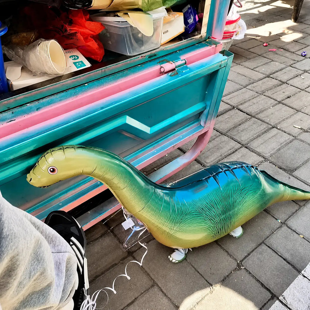 Green dinosaur walking foil balloon displayed outdoors beside a colorful cart