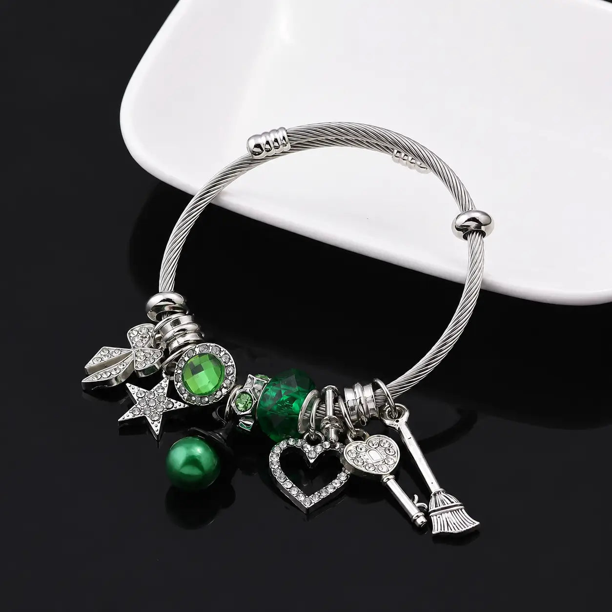 Green and silver witch charm bracelet with heart, star, bow, key, broom, and green bead charms displayed on a black surface.