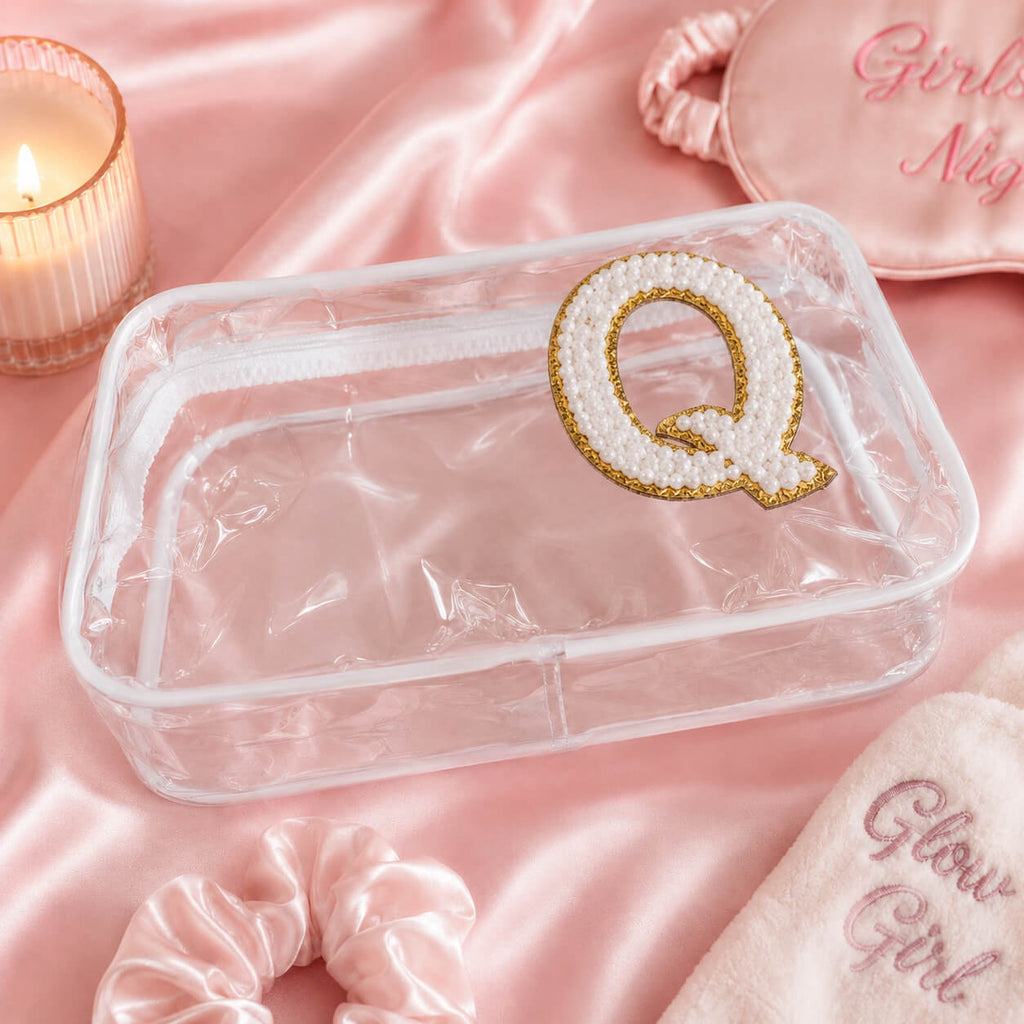 Transparent clear cosmetic pouch with pearl chenille letter Q patch on pink satin background with scrunchie, candle, and sleep mask for girls spa party