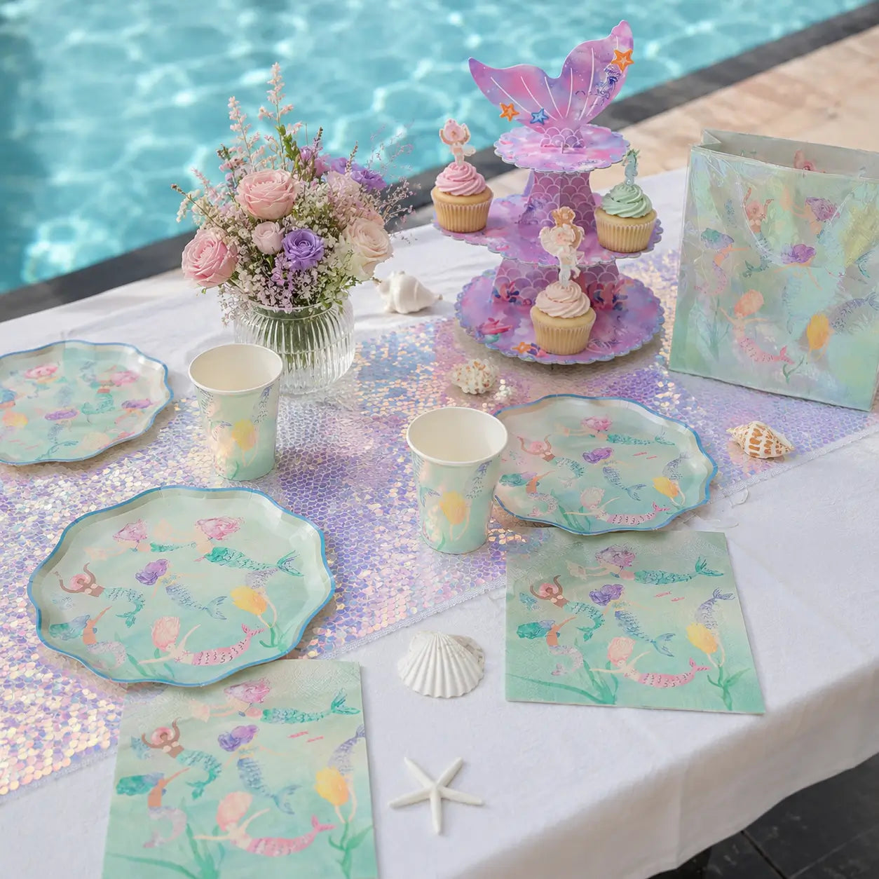Poolside mermaid party table setup with themed plates, cups, napkins, sequin table runner, cake stand and gift bag
