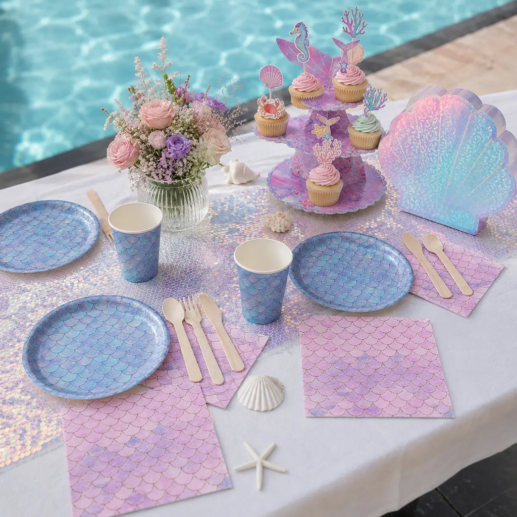 Pastel mermaid pool party table setting with fish-scale tableware, pink napkins, wooden cutlery and an iridescent shell gift bag.