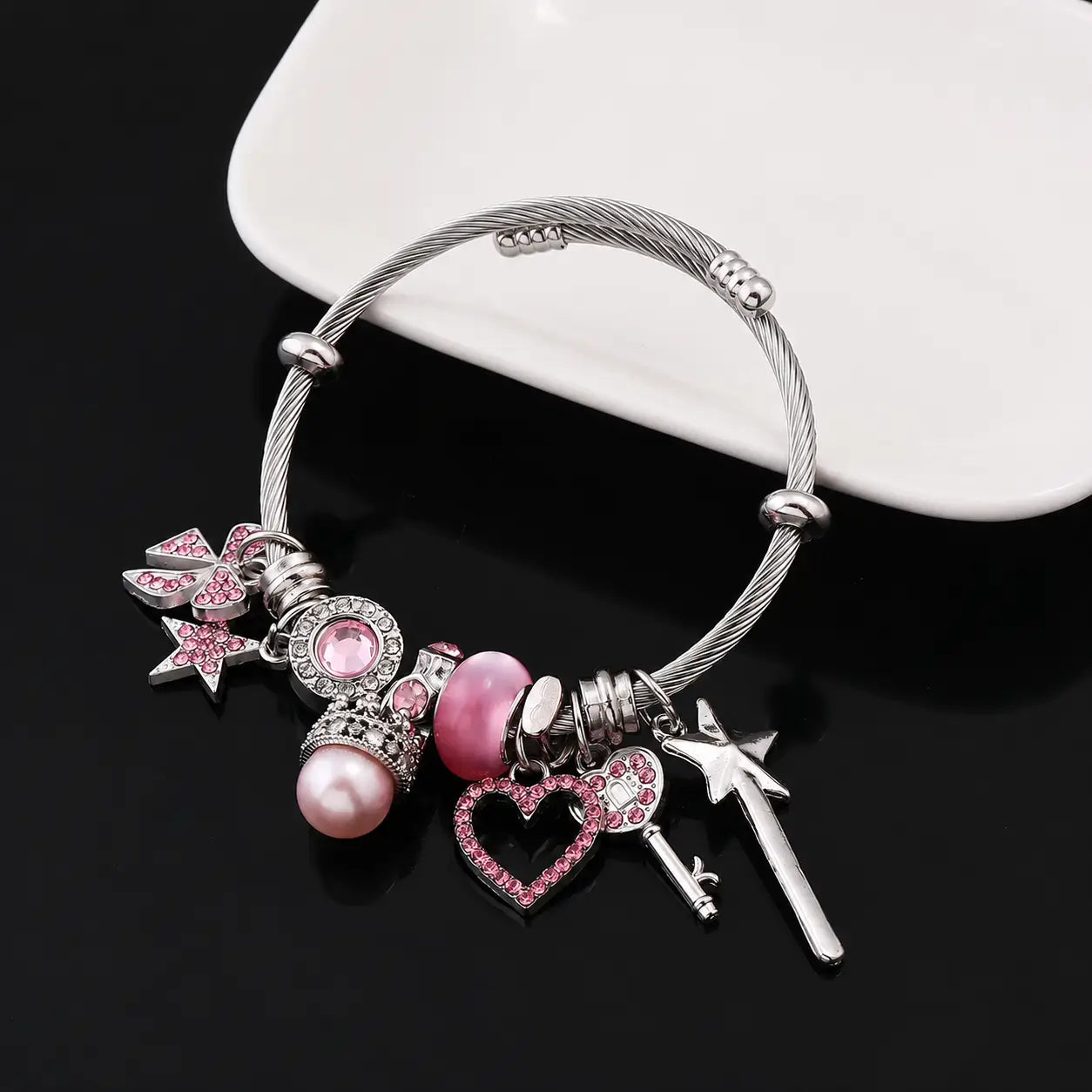 Pink and silver princess charm bracelet with heart, star, bow, key, wand, crown, pearl, and pink rhinestone charms displayed on a black surface.