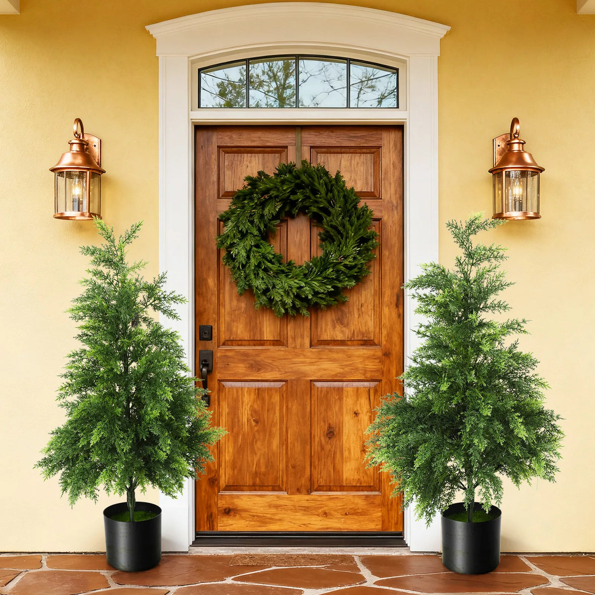 Two potted trees are styled beside a wooden door and a wreath.