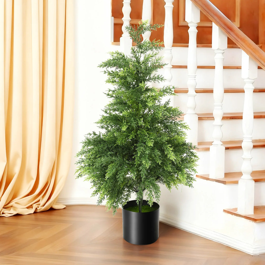 The potted 90 cm Christmas tree stands beside a curtain and wooden staircase.