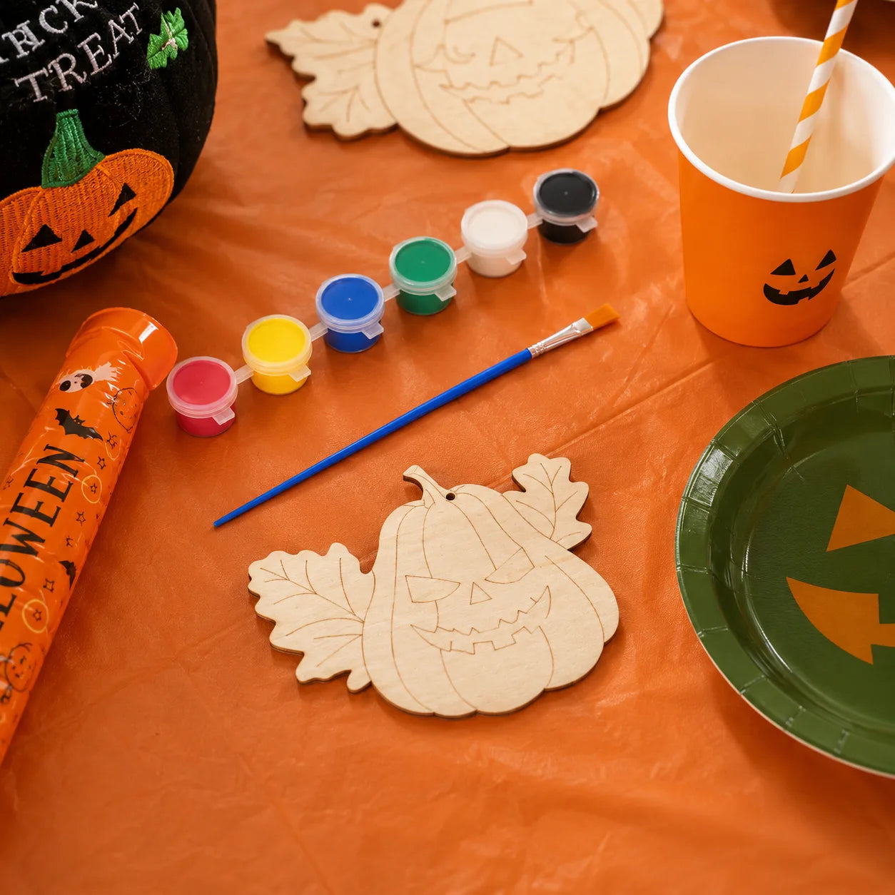 Pumpkin painting craft table with wooden pumpkin ornaments, paint pots, brush, orange cups, pumpkin straws and Halloween tableware