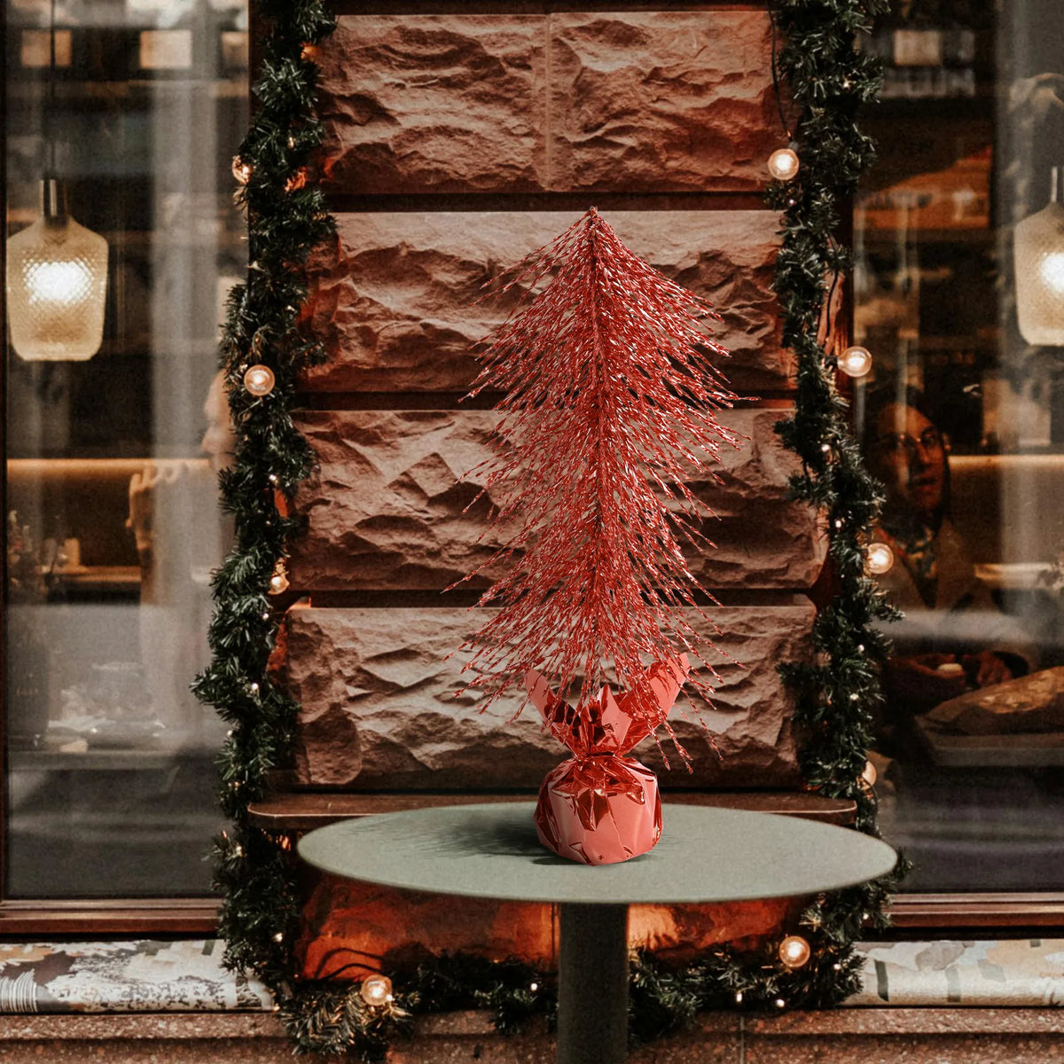 A red tabletop Christmas tree stands on a round table before a fireplace.