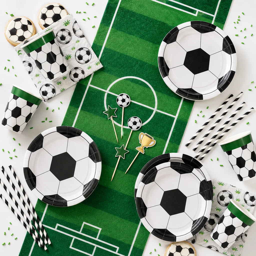 Soccer party tableware flat lay with soccer plates cups napkins striped straws cake toppers and green field table runner