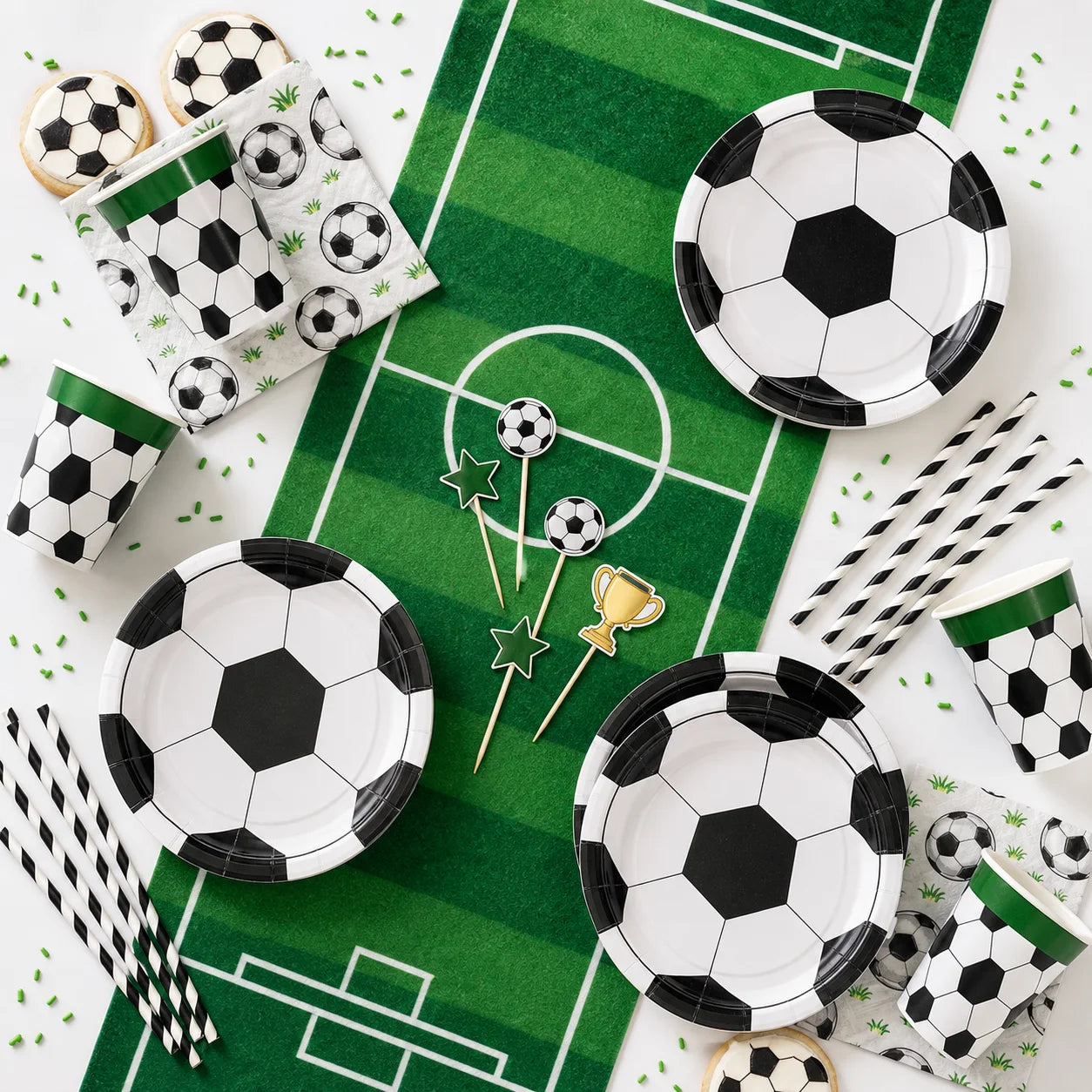 Soccer party tableware flat lay with soccer plates cups napkins striped straws cake toppers and green field table runner