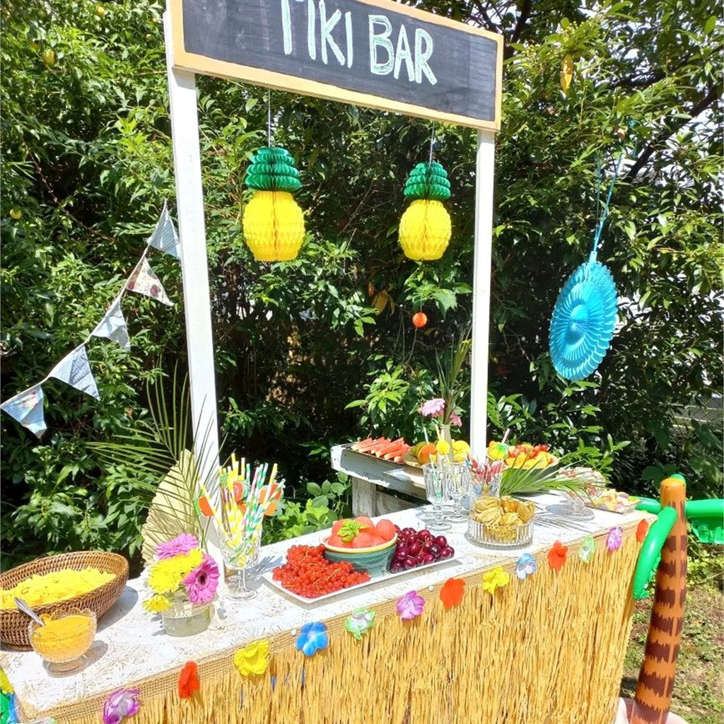 Tiki bar themed summer party buffet with a grass table skirt, hanging pineapples, and colorful straws
