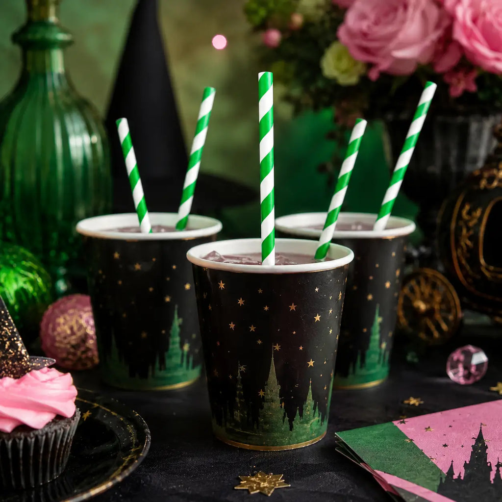 Witch party drink cups with green and white striped paper straws, pink and green napkins, cupcakes, flowers, and magical Halloween table decorations.
