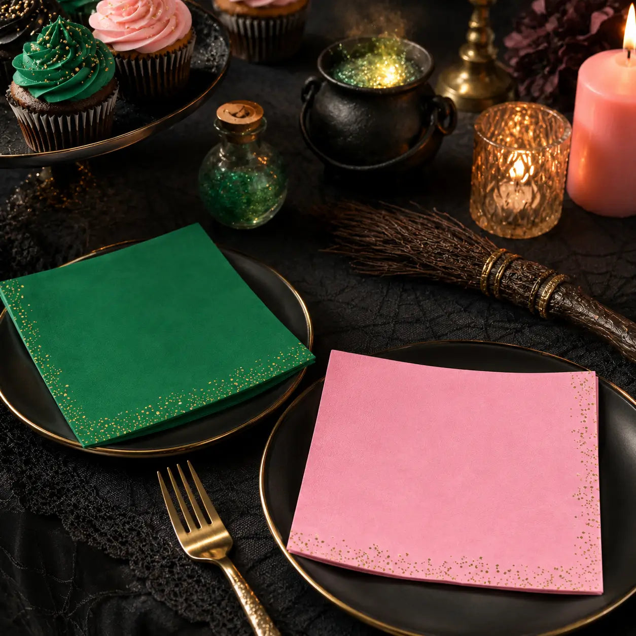 Witch party table setting with green and pink gold glitter napkins, black plates, broom, cauldron, candles, and cupcakes for Halloween party decorations.
